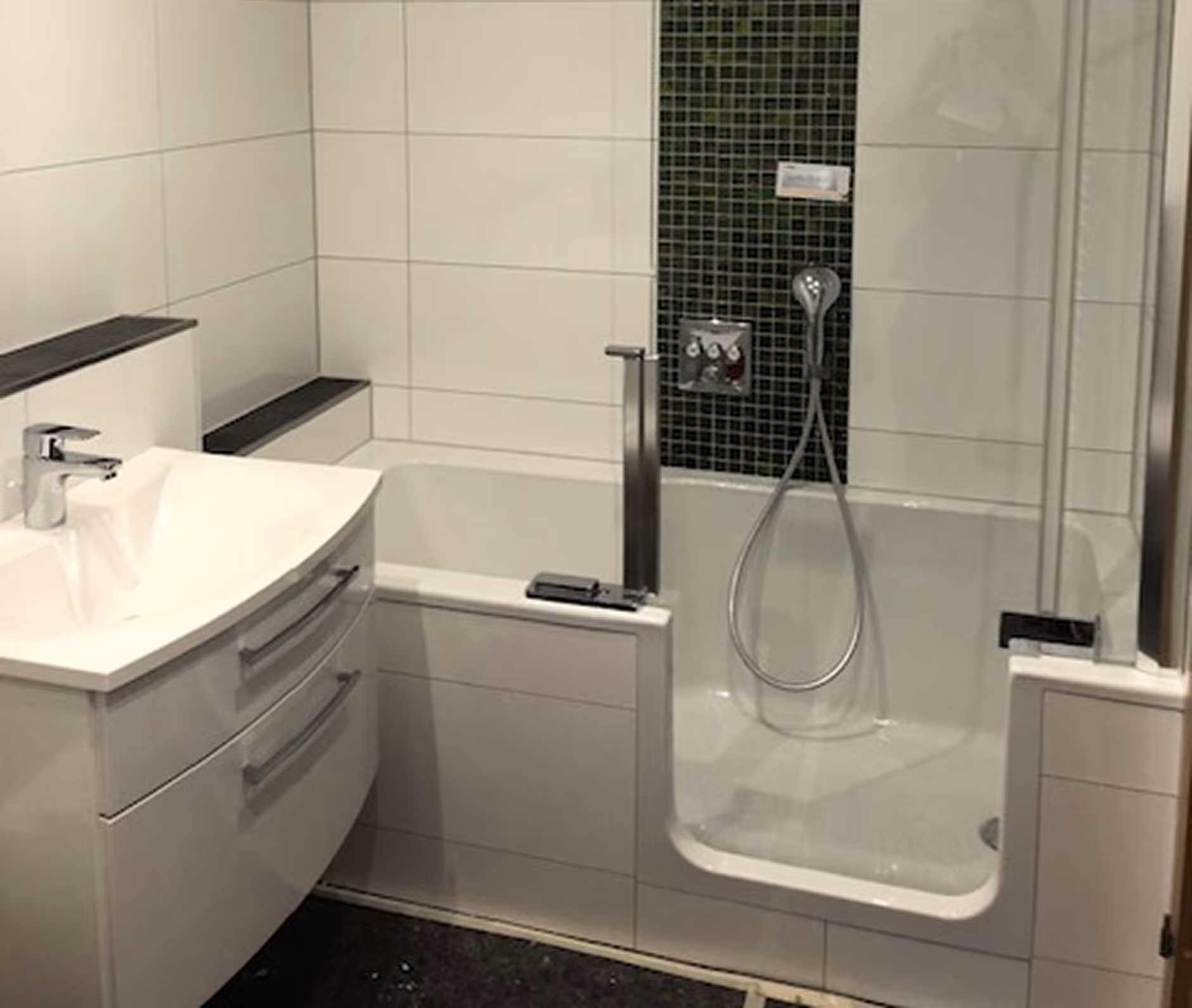 Modernes Badezimmer mit weißem Waschbecken, eingebauter Badewanne mit Glastür und Duschkopf an schwarzer Fliesenwand.