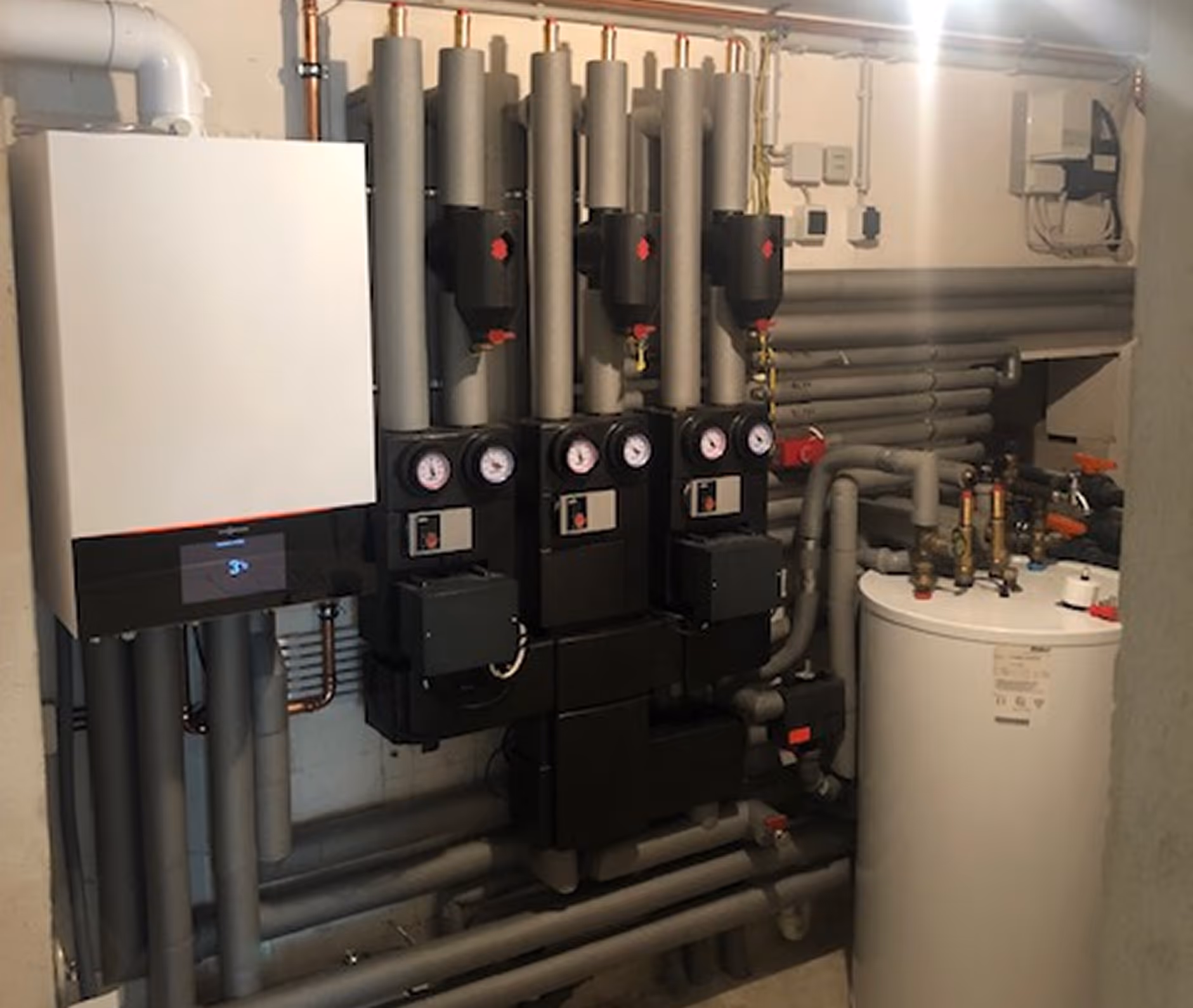 Heizungssystem mit weißem Boiler, mehreren grauen isolierten Rohren, Messgeräten und Kontrollkästen in einem Hauswirtschaftsraum.