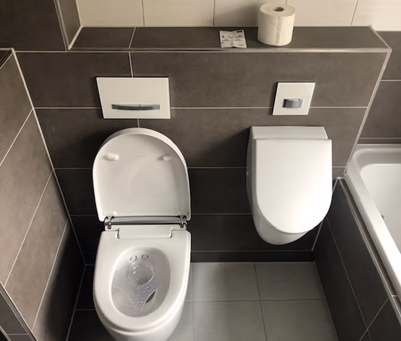 Moderne Toilette mit geöffnetem Sitz und geschlossenem Deckel an gefliester Wand, darüber eine Rolle Toilettenpapier.