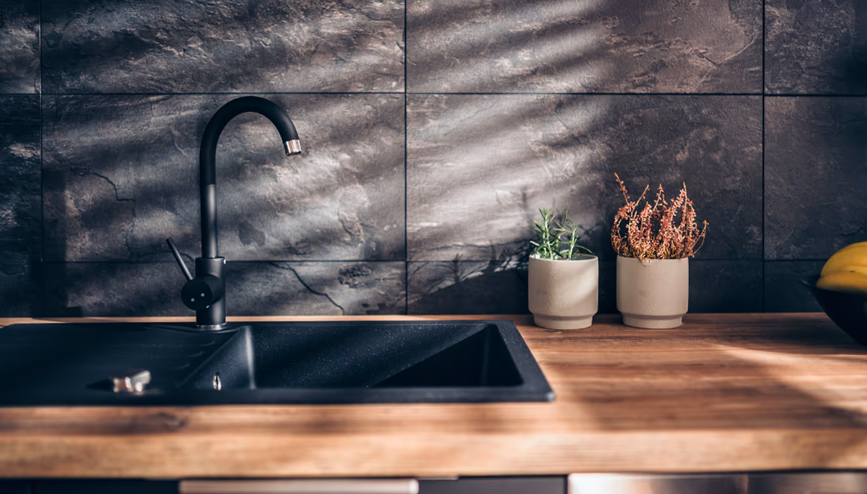 Moderne Küche mit schwarzer Spüle und Wasserhahn, zwei kleinen Pflanzentöpfen und Holzarbeitsplatte vor dunkler Steinfliesen-Wand.