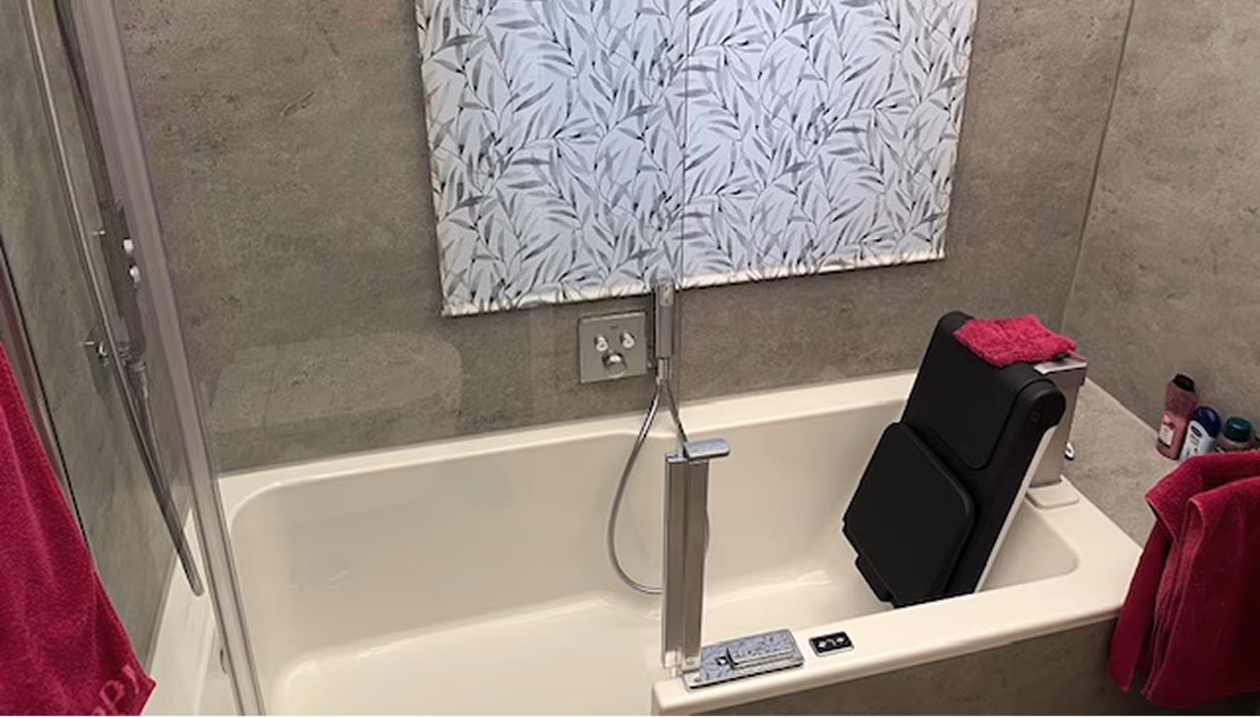 Badewanne mit verstellbarem Sitz, grauen Fliesen und rosa Handtüchern.