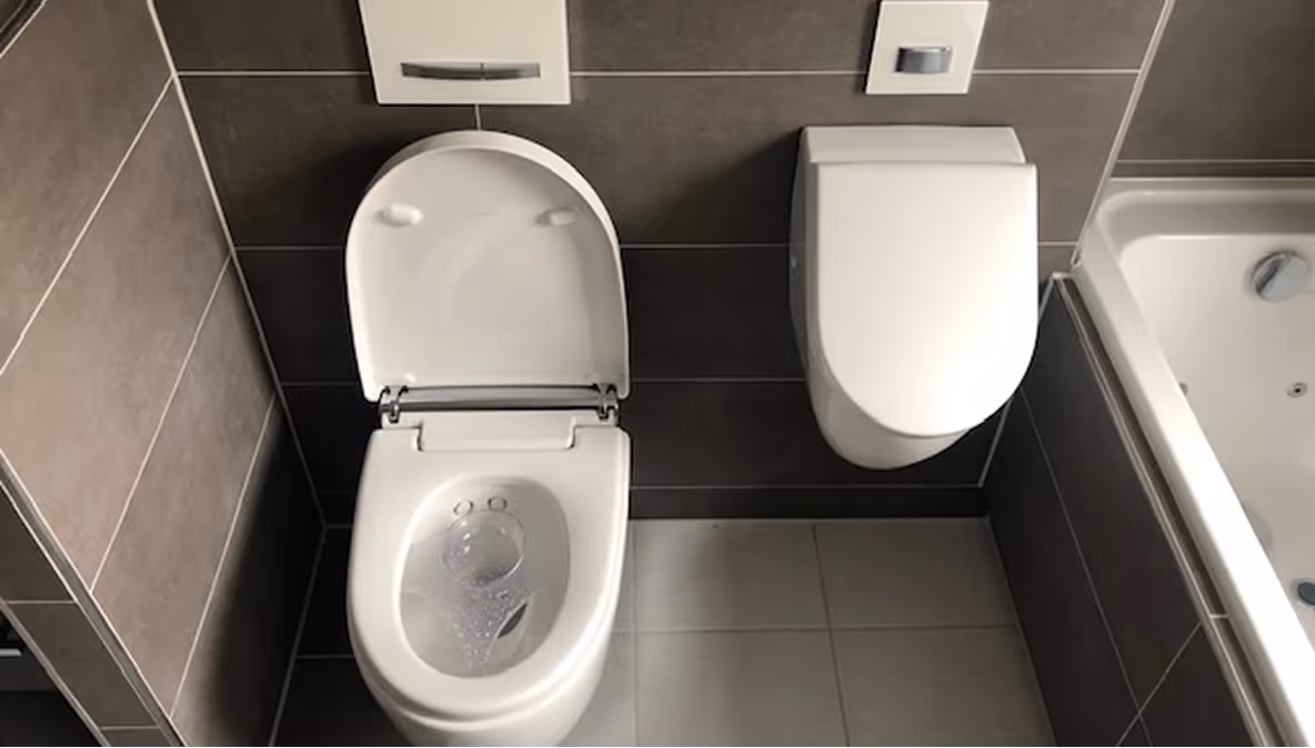 Moderne Toilette mit offenem Sitz und geschlossenem Deckel an einer gefliesten Wand neben einer Badewanne.