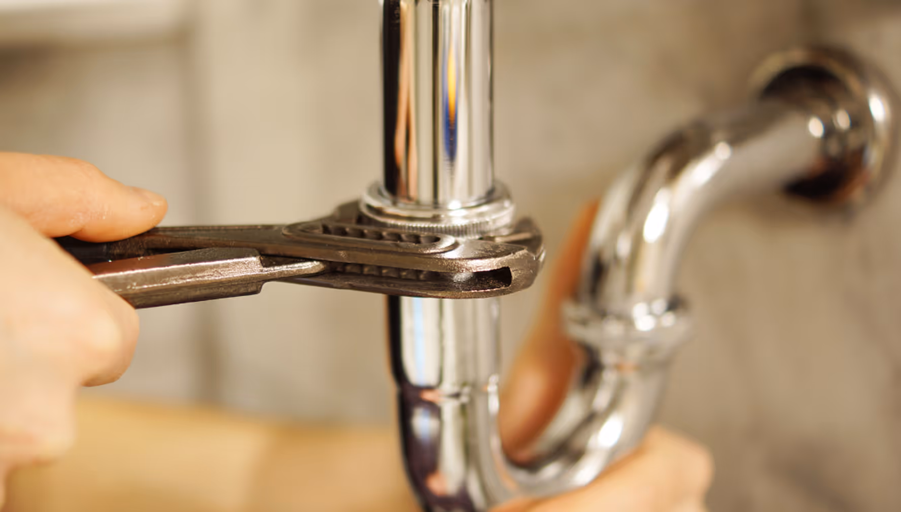 Hand hält Rohrzange und repariert verchromtes Abflussrohr unter einem Waschbecken.