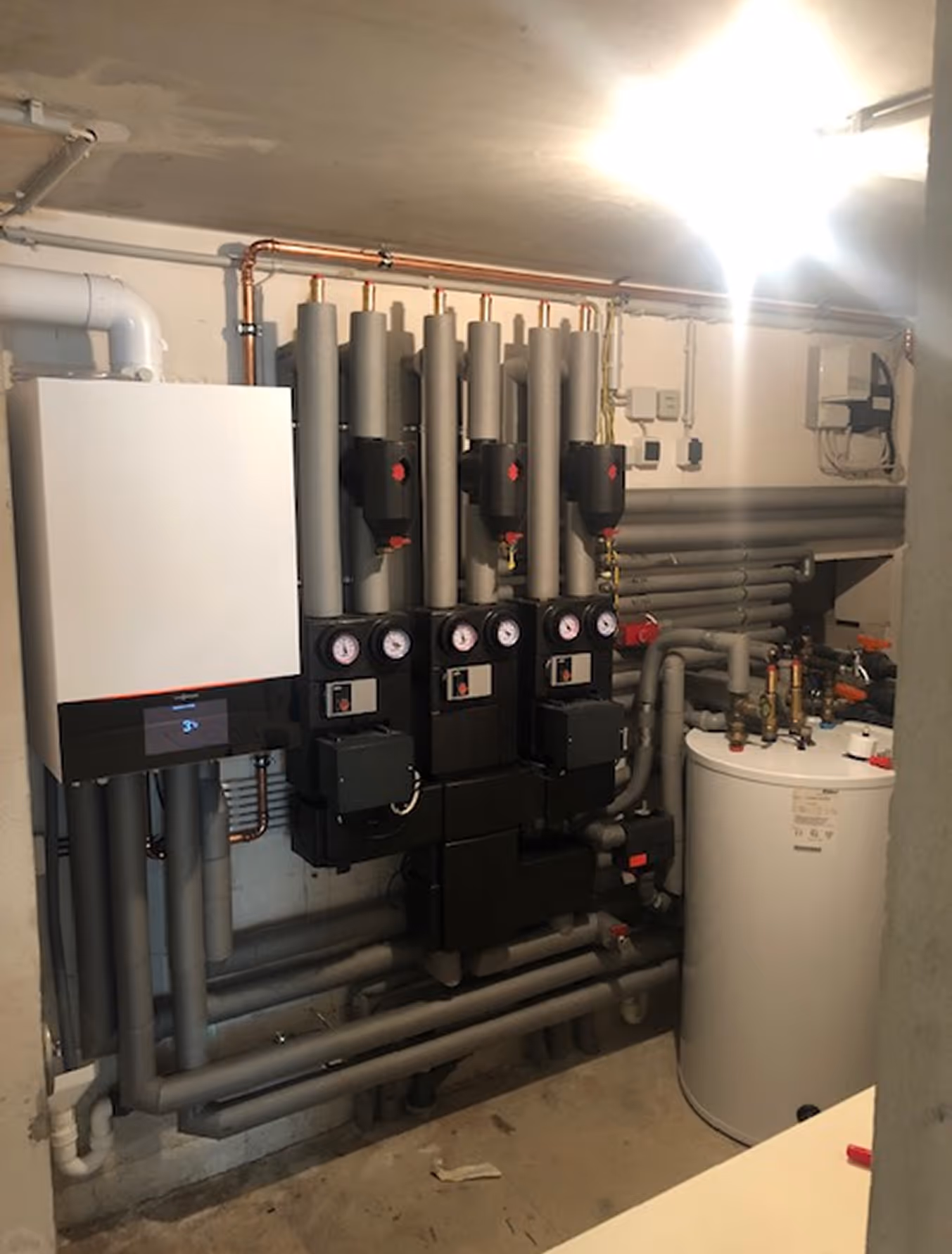 Heizungsanlage mit Rohren, Kontrollanzeigen und einem weißen Warmwasserspeicher in einem Kellerraum.