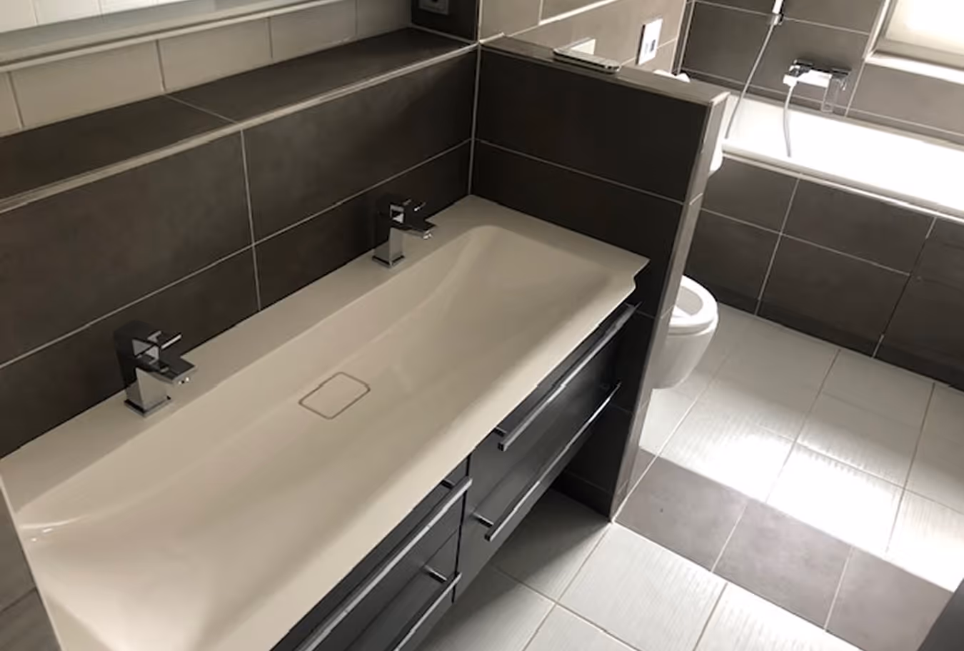 Modernes Badezimmer mit weißem Doppelwaschbecken, grauen Schränken, einer Toilette und einer Badewanne neben dem Fenster.