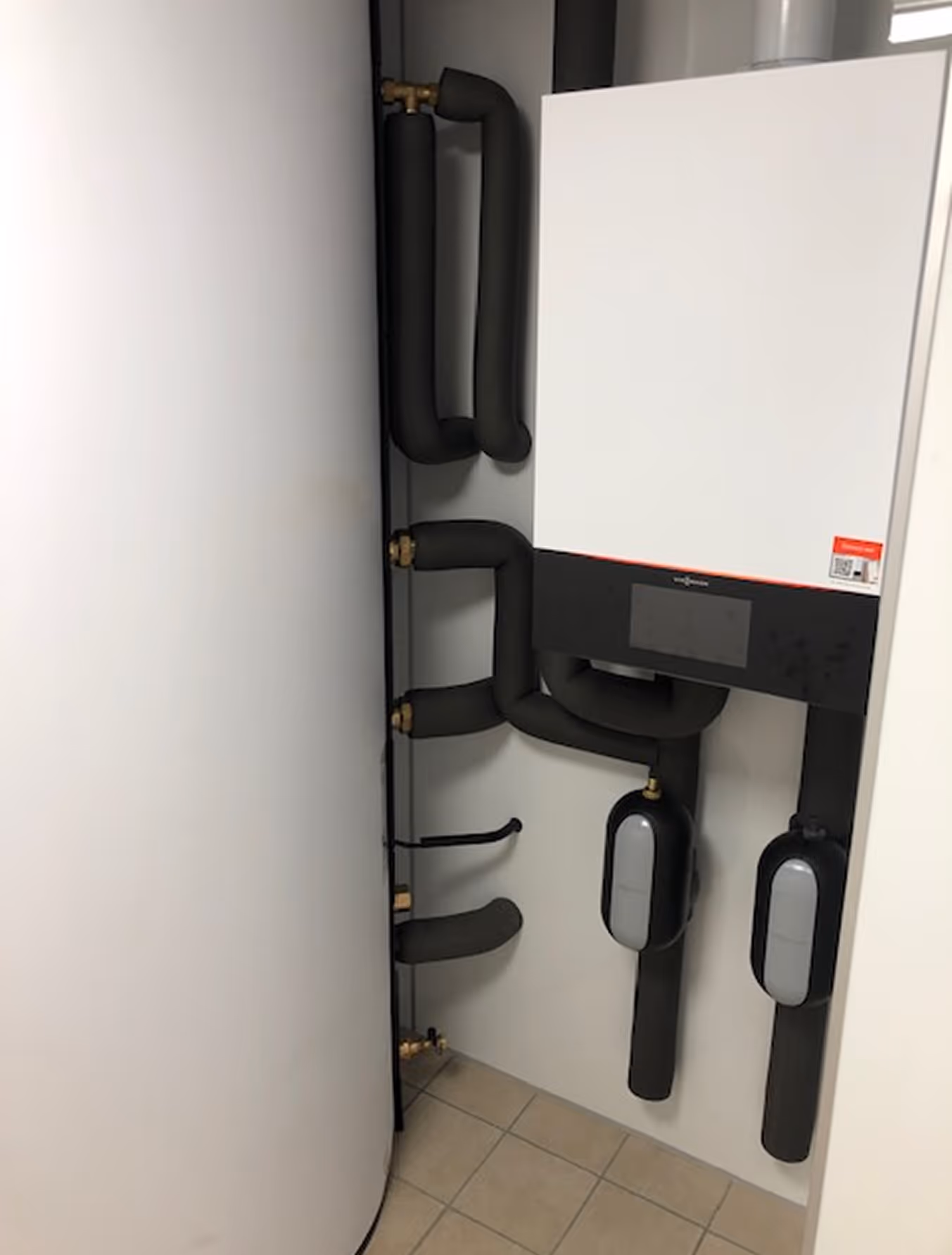 Weiße Heizungsanlage mit schwarzen isolierten Rohren in einem Raum mit Fliesenboden.