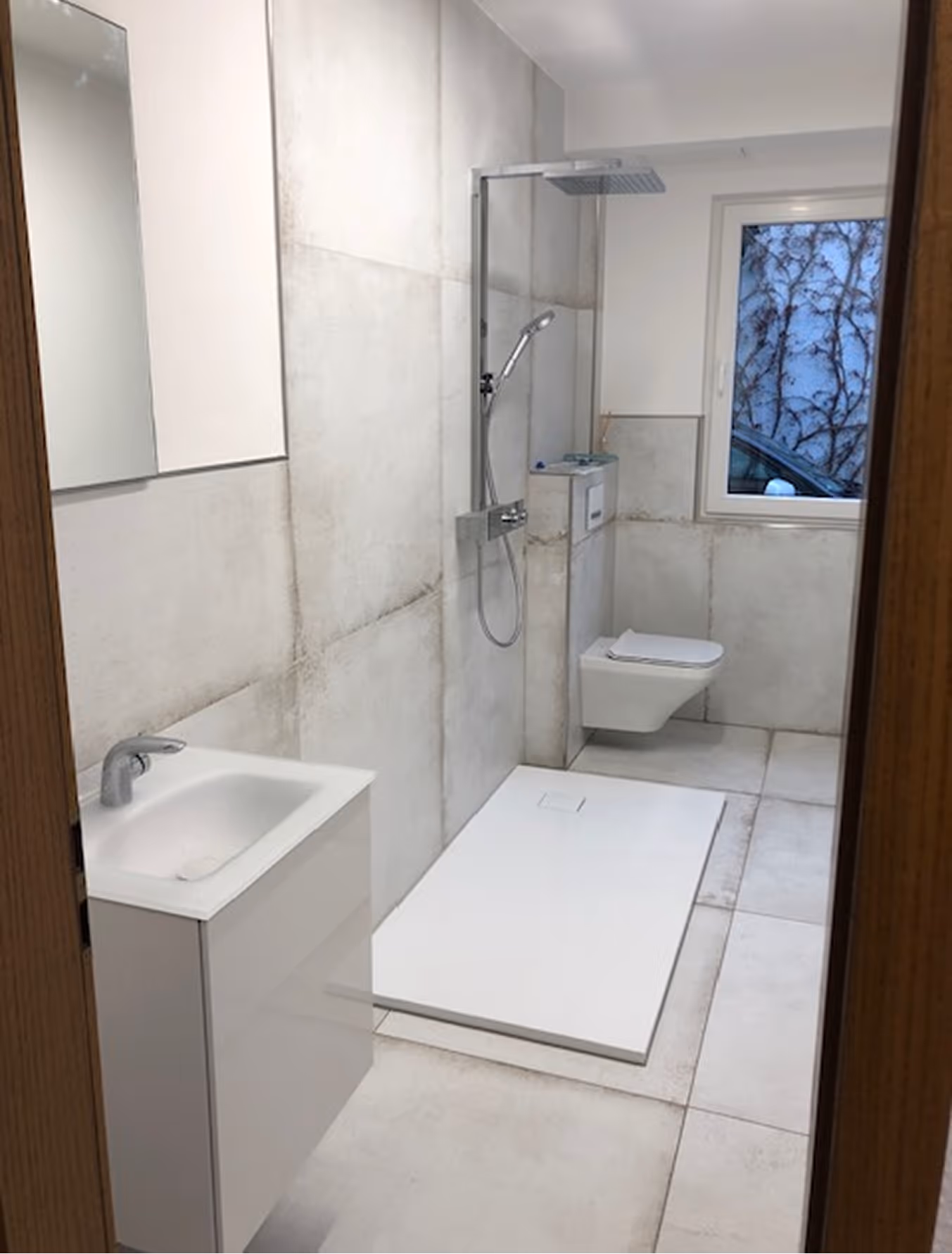 Modernes Badezimmer mit weißem Waschbecken, bodengleicher Dusche und wandhängendem WC unter einem Fenster.